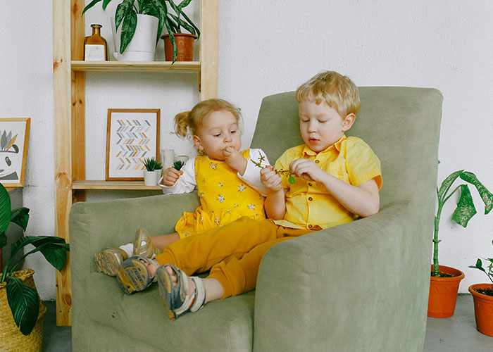 Two young children in yellow outfits sitting on a green chair surrounded by plants in a cozy living room setting