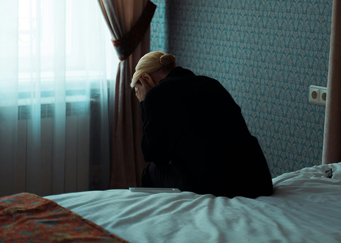 Woman sitting on bed with head in hands in dimly lit room, illustrating stress of traditional wives’ real experiences.