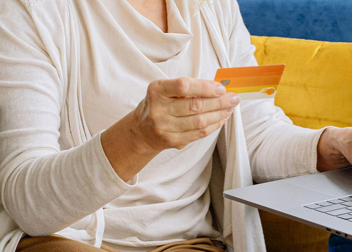 Middle-aged woman holding a credit card while using a laptop, representing traditional wives managing finances in the 20th century.