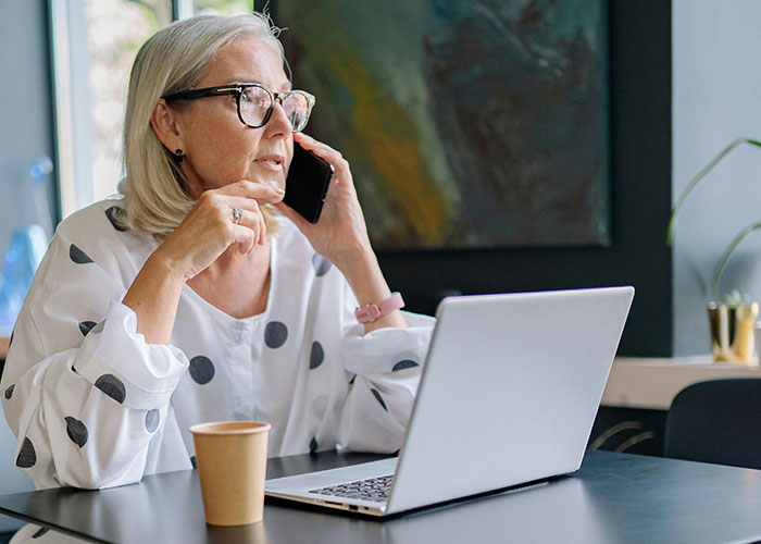 Older woman in glasses talking on phone while working on laptop, reflecting on traditional wives 20th century experiences.