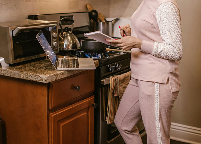 Traditional wife in 20th century kitchen multitasking with laptop and notepad near stove and microwave oven.