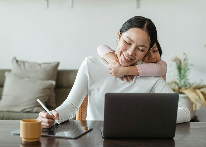 Woman working on a laptop while her child hugs her, illustrating traditional wives balancing work and family life.