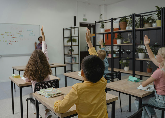 Classroom scene with children raising hands while a teacher shares stories about traditional wives' real experiences.