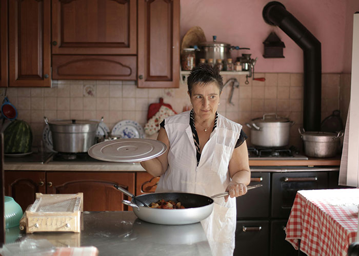 Traditional wife in a 20th century kitchen cooking on stove, wearing apron and holding pot lid, preparing a meal.