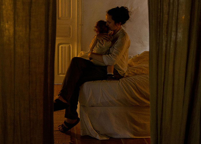 Traditional wife from the 20th century sitting on bed holding child in a dimly lit room, sharing real experiences.