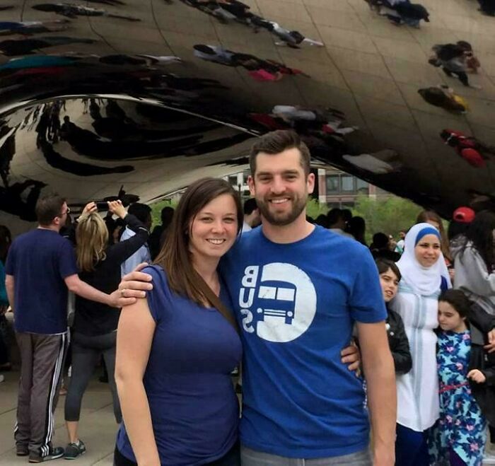 Couple posing in front of a reflective sculpture creating a confusing optical illusion effect, with people mirrored in background.
