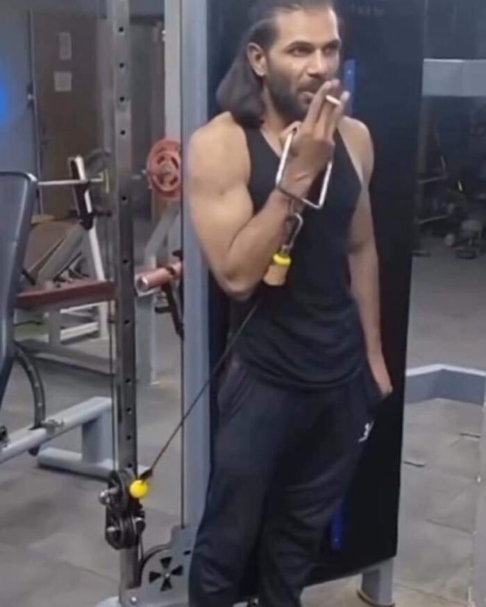 Man in a gym smoking, uses a cable machine creatively, showcasing a blend of humor and irony.