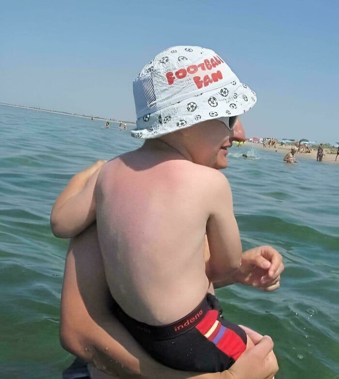 Man and child at beach, optical illusion makes arms appear confusingly intertwined, wearing a "Football Fan" hat.