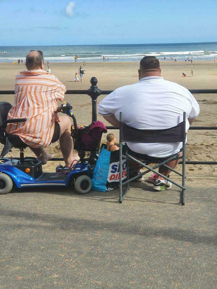 Two individuals sitting on a beach, creating an optical illusion with a doll in a bag, causing a double take.