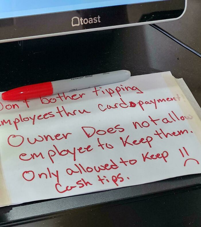 Note about tipping policy written in red marker on paper at a restaurant register, illustrating ridiculous capitalism issues.