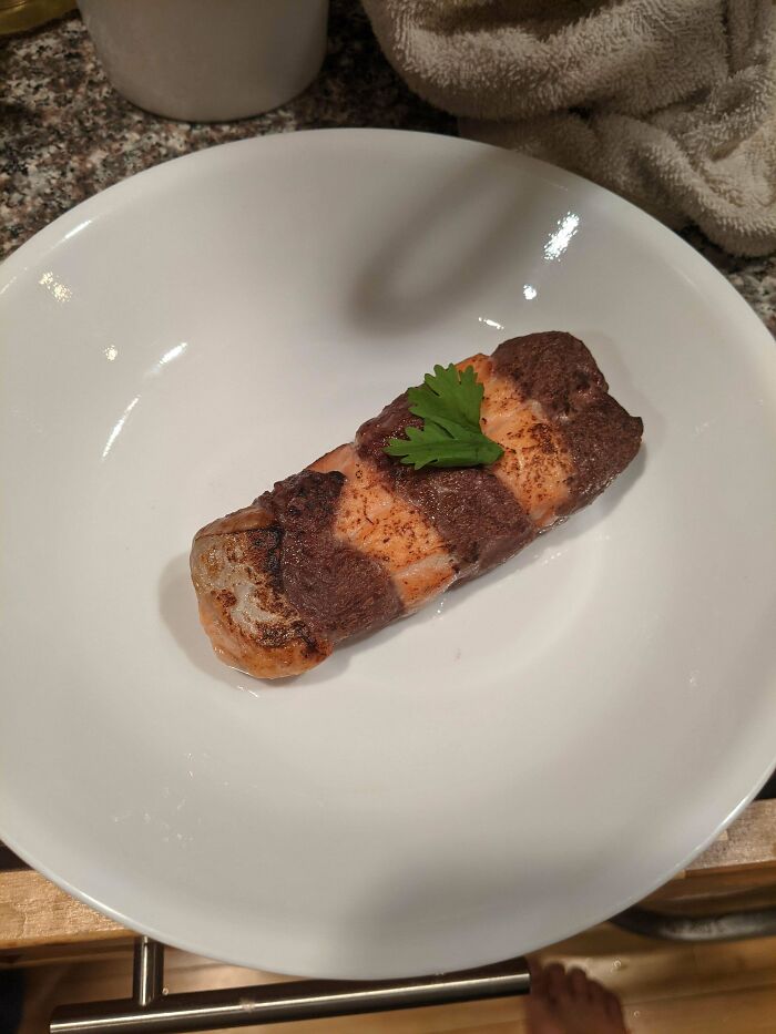 A bizarre food creation featuring a tortilla-wrapped salmon roll garnished with a cilantro leaf, presented in a white dish.