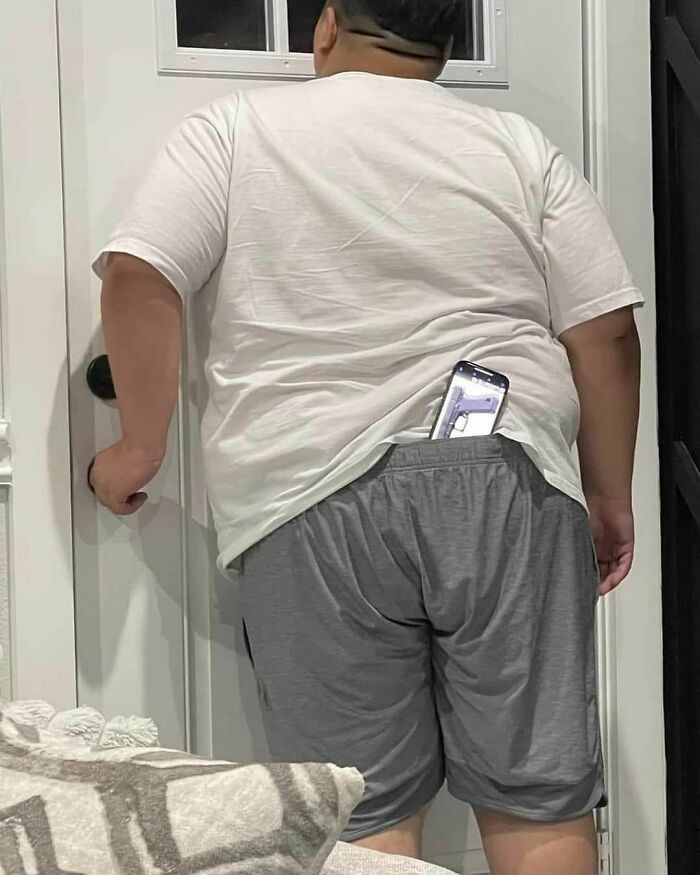 Person wearing a white shirt and gray shorts stands at a door, phone tucked into their waistband.