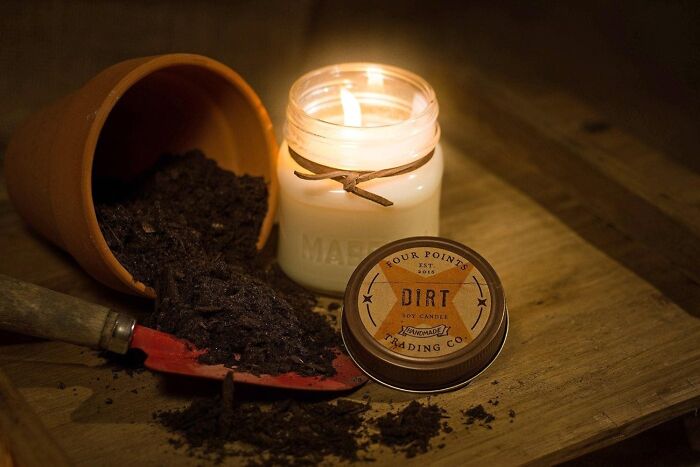 Fallen plant pot with soil, a lit candle labeled "Dirt" creating a cozy ambiance on a wooden surface.