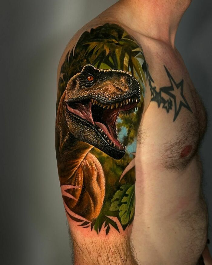 A detailed dinosaur tattoo featuring a roaring T-Rex on a man's upper arm, surrounded by jungle foliage.