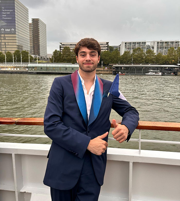 French diver on a boat, wearing a blue suit with a colorful lapel, standing with a thumbs up by the river.