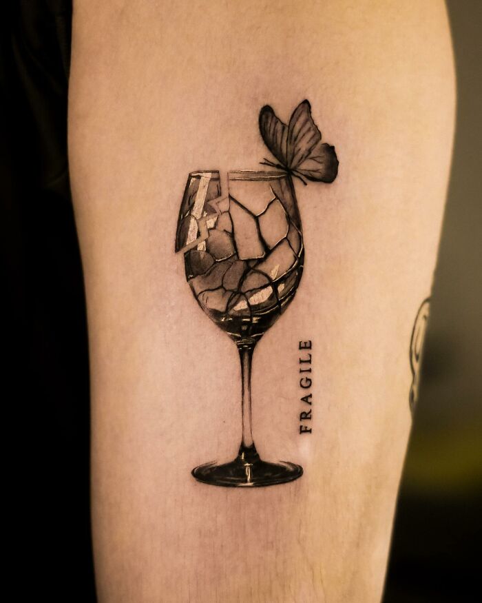 Hyperrealistic tattoo of a shattered wine glass with a butterfly perched on it, labeled "Fragile."