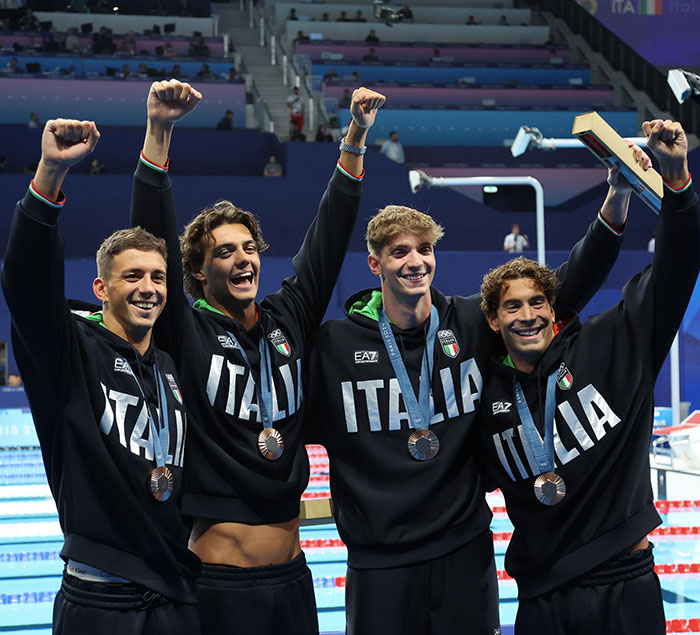 Four Italian swimmers celebrate with medals around their necks, cheering on the podium.