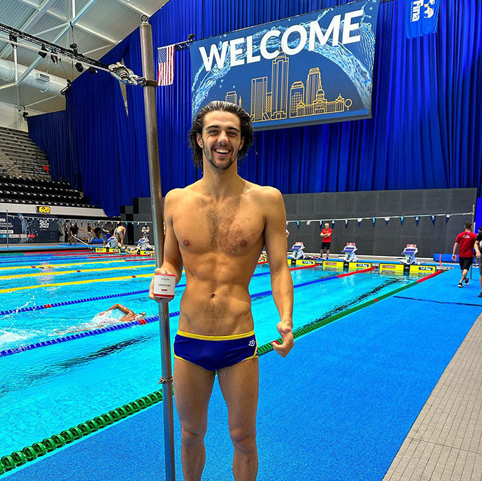 Gold medalist in swim briefs at a pool event, smiling and holding onto a pole, with a blue backdrop and welcome sign.