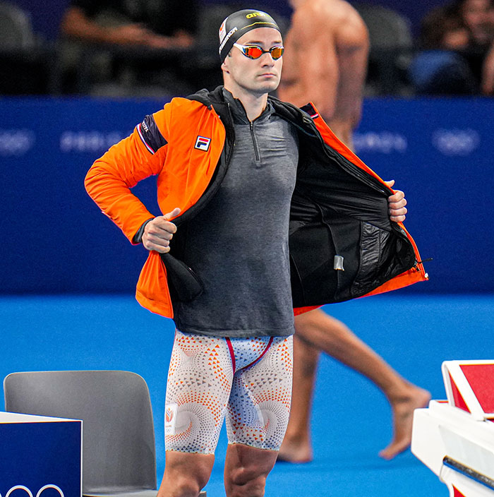 Dutch swimmer in revealing trunks at the 2024 Olympics poolside, wearing an orange jacket and swimming cap.