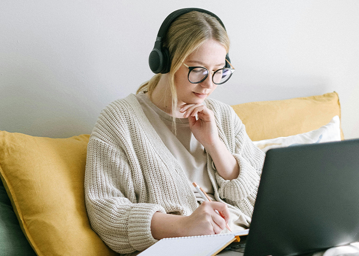 Young woman with headphones studying slang for Gen Alpha at a laptop, writing notes.