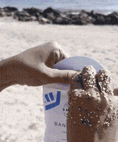 Hands covered in sand holding a white TikTok Shop product pouch on a sunny beach with rocks in the background.