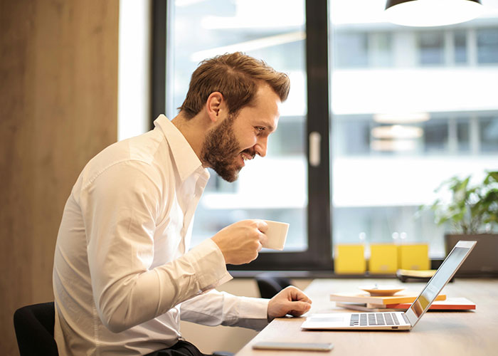 Man in a white shirt smiling at laptop screen while holding a coffee cup, illustrating work mess-ups and challenges.