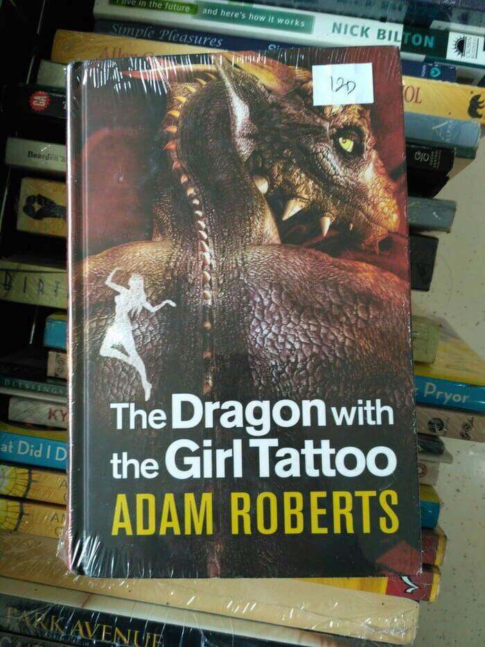 Book titled The Dragon with the Girl Tattoo by Adam Roberts, shown among a stack of various books, showcasing brand rip-offs.