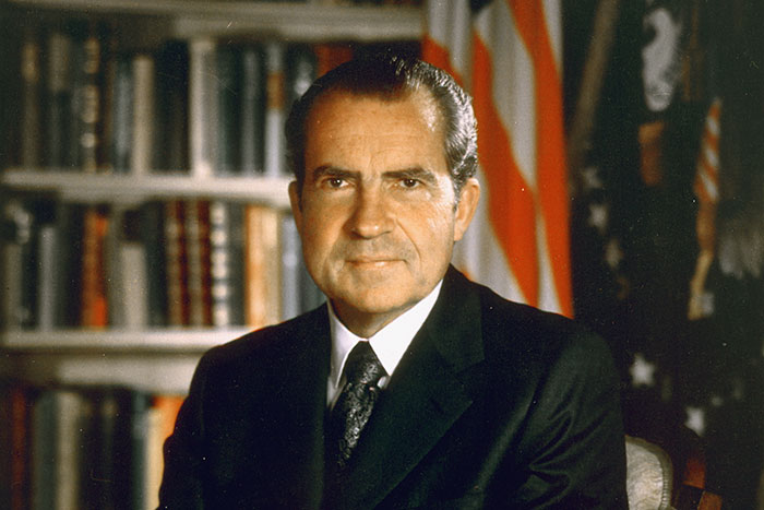 Portrait of a historical figure in a suit with bookshelves and a flag in the background.