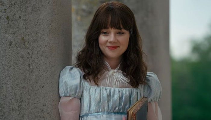 Makeup artist covers up tattoo on actress Claudia Jessie in period dress, standing against a stone pillar, looking content.