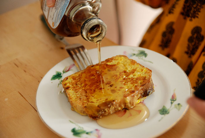 Pouring syrup on a slice of French toast on a floral plate, illustrating food stereotypes about people’s home countries.