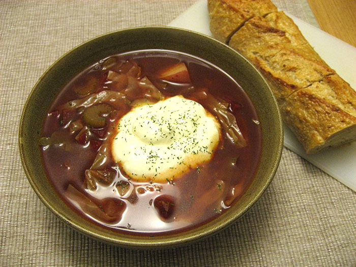 Bowl of traditional soup with sour cream and herbs next to sliced crusty bread representing home country stereotypes.