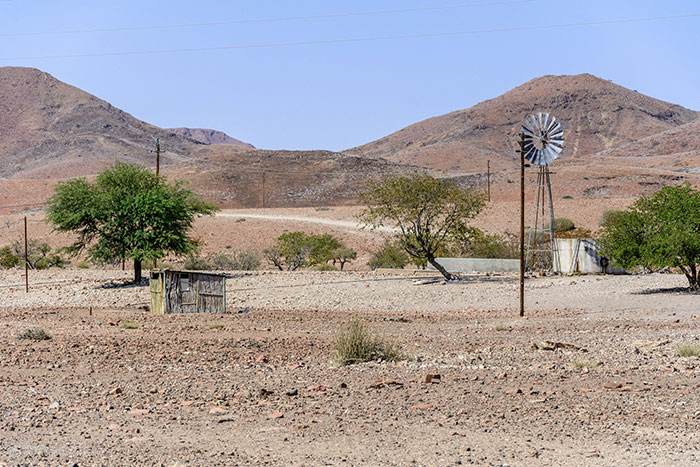 Arid rural landscape with sparse trees, a windmill, and mountains, reflecting stereotypes about home countries.