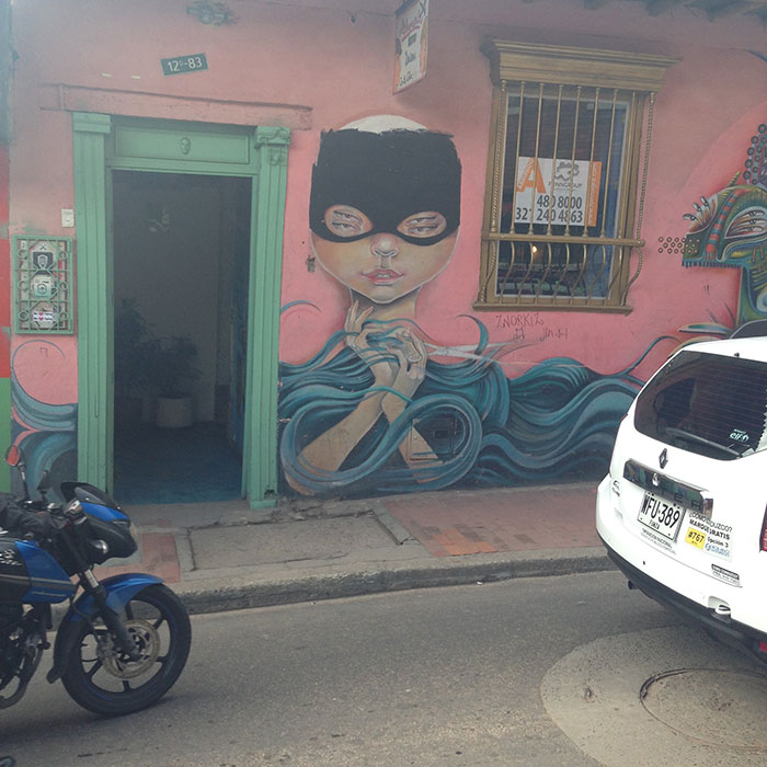 Street mural of a masked figure with flowing hair on a pink wall, parked motorcycle and white car nearby in urban setting.