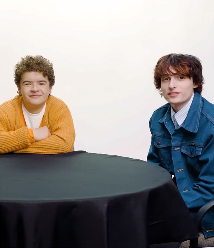 "Stranger Things stars seated at a table, one in an orange sweater, the other in a denim jacket, smiling at the camera. "Stranger Things stars seated at a table, one in an orange sweater, the other in a denim jacket, smiling at the camera.