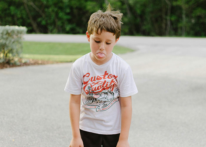 Young boy sticking out tongue, outdoors on a paved driveway, illustrating themes of unethical parenting hacks and manipulation.