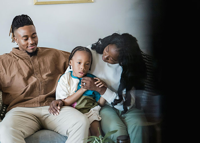 A family of parents sitting with their child on a couch, illustrating unethical parenting hacks and manipulation techniques.