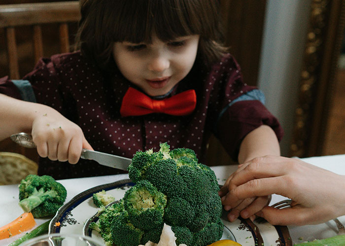 Child using a knife to cut broccoli at the table, illustrating unethical parenting hacks involving food manipulation.