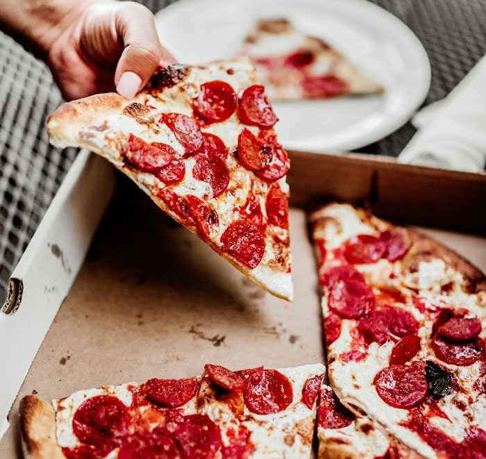 Hand holding a slice of pepperoni pizza from a box, illustrating lifetime supply winnings of pizza experiences.