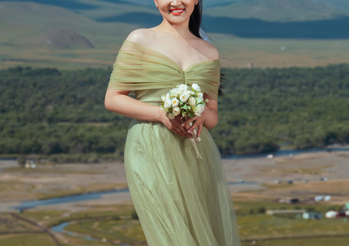 Bride in a green dress holding a bouquet, standing outdoors with a scenic landscape in the background.