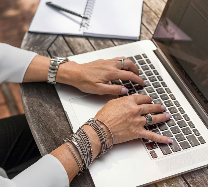 Person typing on laptop outdoors with multiple bracelets and rings, illustrating embarrassing attempts at understanding computers.