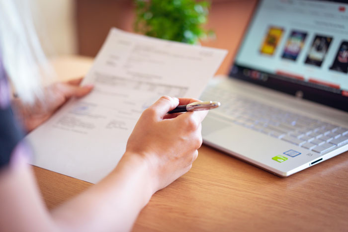 Person holding a pen and document, sitting at a desk with a laptop, illustrating embarrassing attempts at understanding computers.