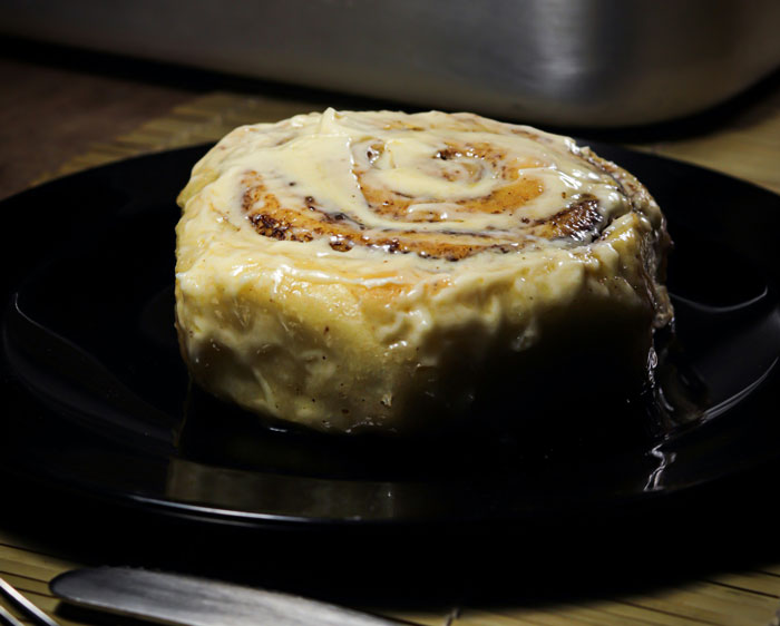 Close-up of a cinnamon roll with creamy icing on a black plate, representing lifetime supply winnings of baked goods.