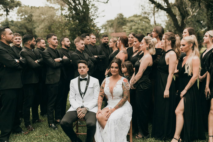Bride and groom sitting, surrounded by bridesmaids and groomsmen in black attire, outdoors.