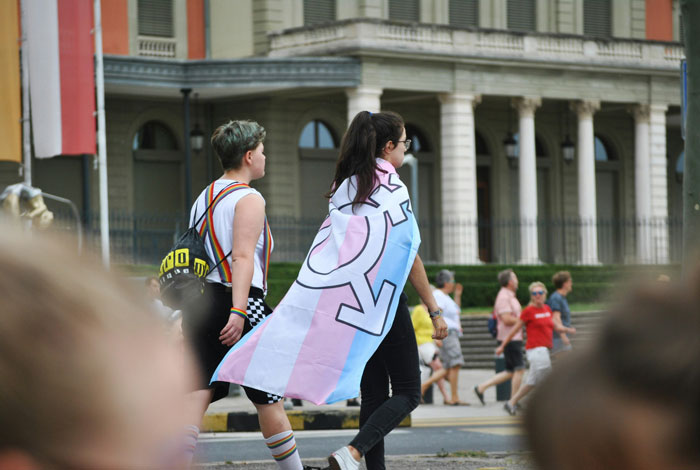 People walking, one wearing a trans flag cape, in front of a historic building, representing inclusivity and diversity.