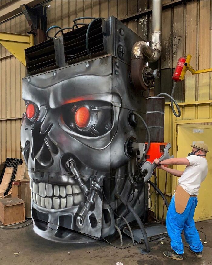 Artist from France working on a giant 3D graffiti with a robotic skull design on industrial equipment indoors.