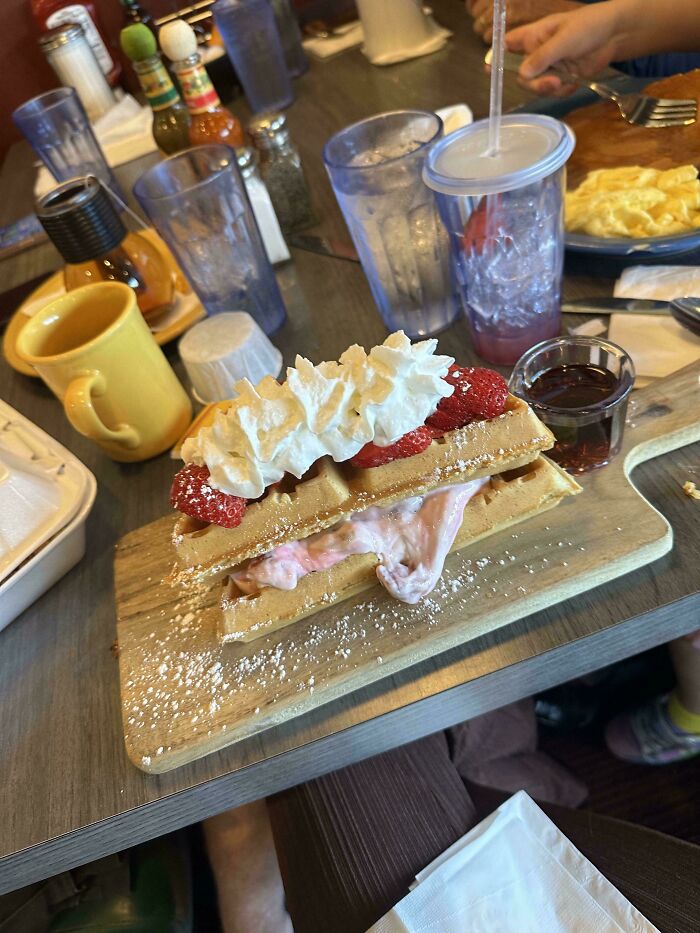 Waffles served with strawberries and whipped cream on a wooden board among drinks and breakfast items.