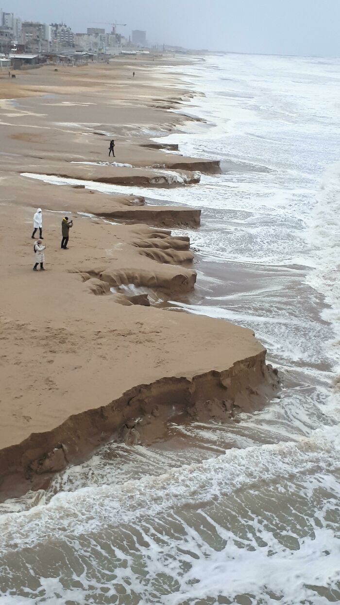 Eroded sandy coastline with people standing near dramatic cliffs, showcasing nature's powerful forces on the shore.