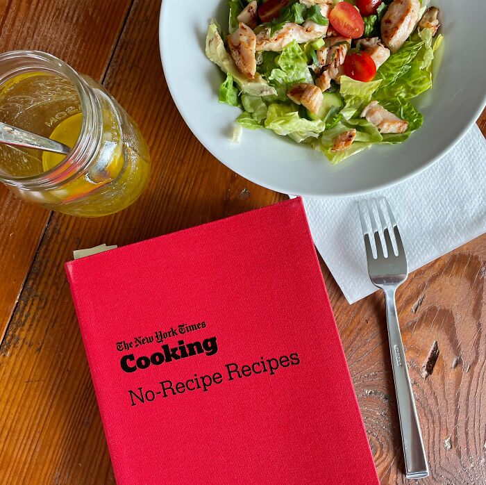 Red cookbook with salad bowl and drink mason jar on wooden table.