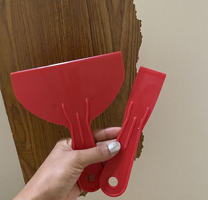 Hand holding red plastic scrapers used for life hacks on damaged wooden wall surface for quick repair.