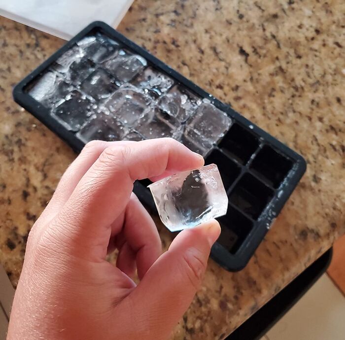Hand holding a clear ice cube above a frozen ice tray on a kitchen counter showing simple life hacks.